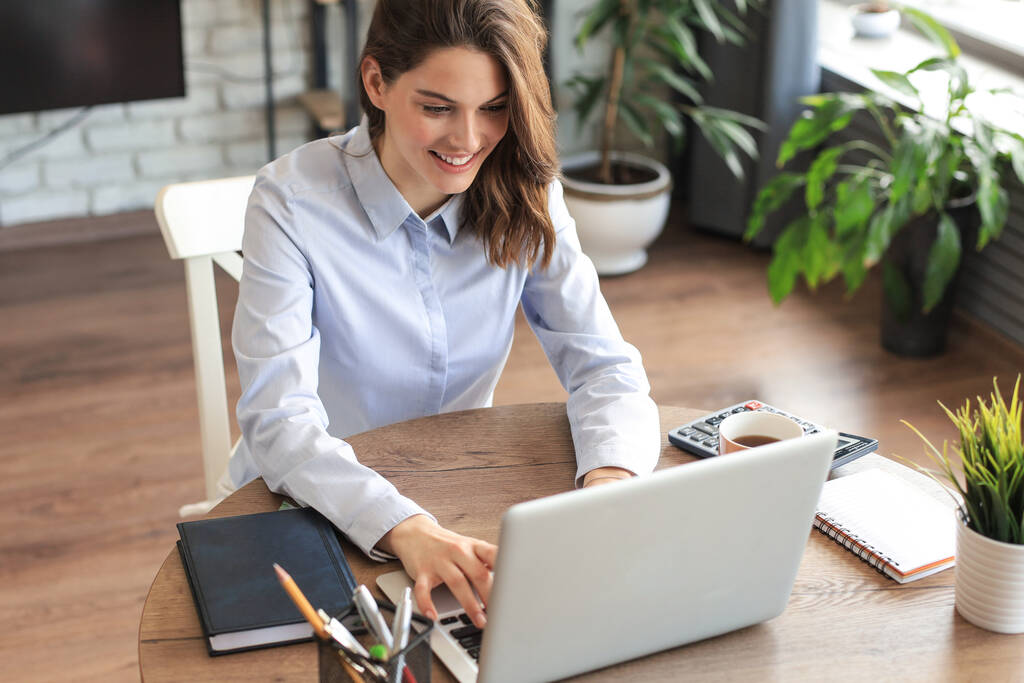 Smiling pretty woman sitting at table, looking at laptop screen. Happy entrepreneur reading message email with good news, chatting with clients online - Photo, Image
