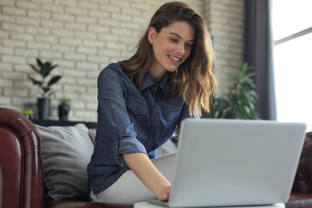 Kanepede dizüstü bilgisayarla oturan ve arkadaşlarıyla sohbet eden gülümseyen genç bir kadın. - Fotoğraf, Görsel