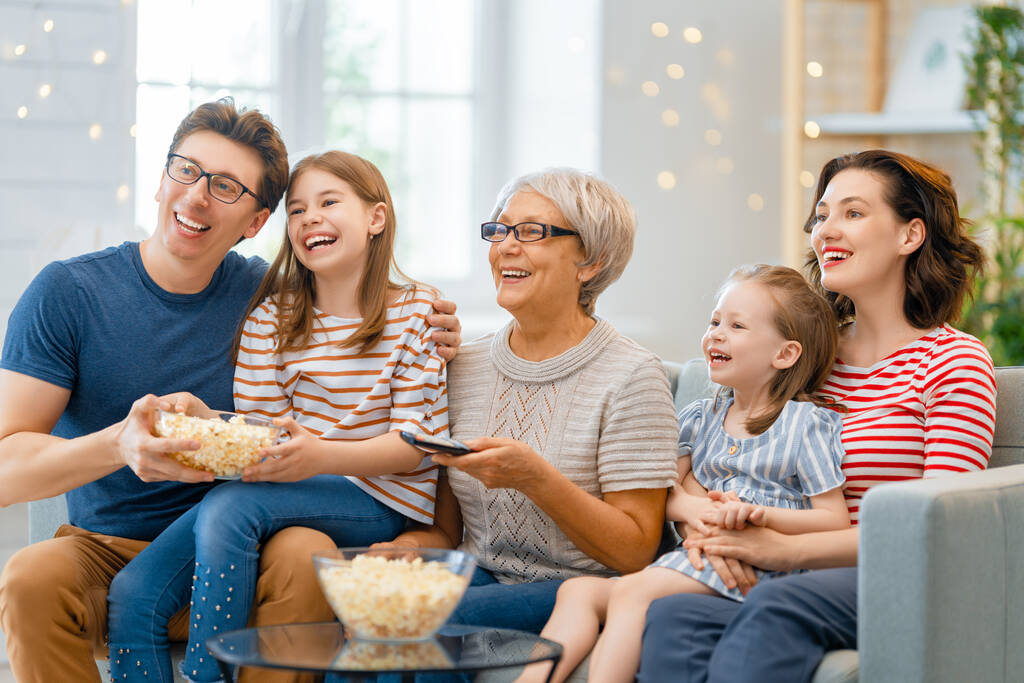 Happy family watching TV with popcorn  at home. Granny, mother, father and daughters spending time together. - Photo, Image