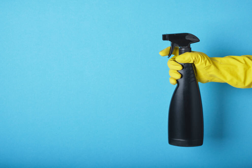 Una mano en un guante amarillo sostiene un aerosol de líquido de limpieza sobre un espacio de copia de fondo azul - Foto, imagen