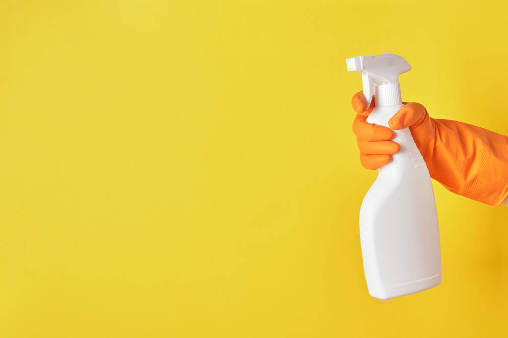 Eine Hand in einem orangefarbenen Handschuh hält eine weiße Sprühflasche mit Reinigungsflüssigkeit auf gelbem Hintergrund. - Foto, Bild