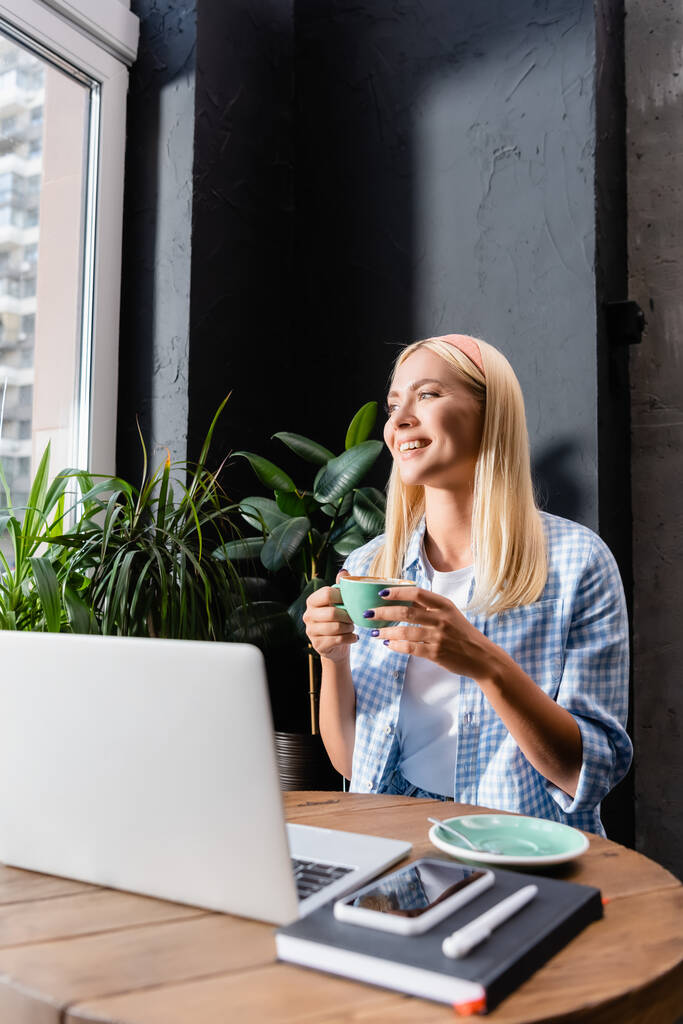 feliz freelancer sosteniendo taza de café cerca de la computadora portátil en primer plano borrosa - Foto, imagen