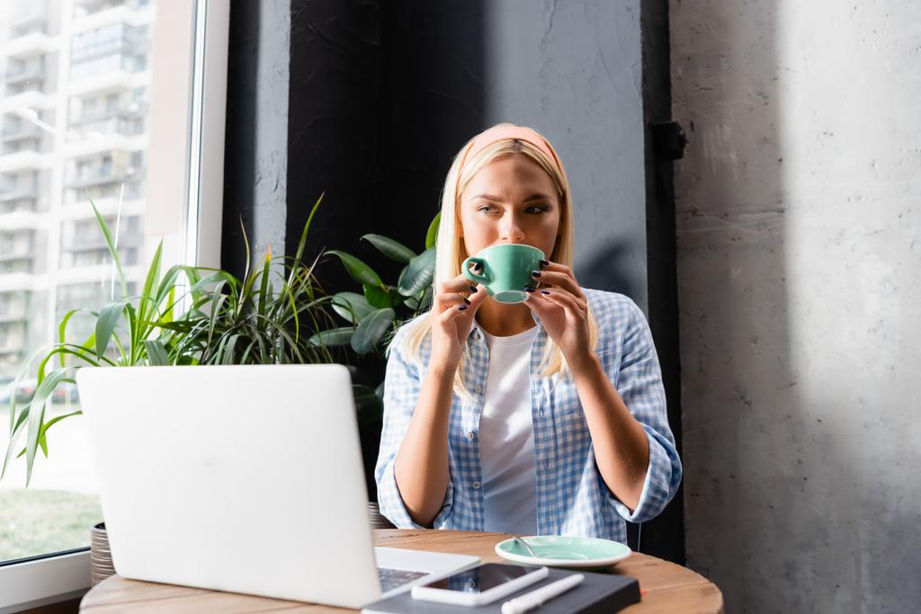 freelance loira bebendo café perto de laptop no café, foreground borrado - Foto, Imagem
