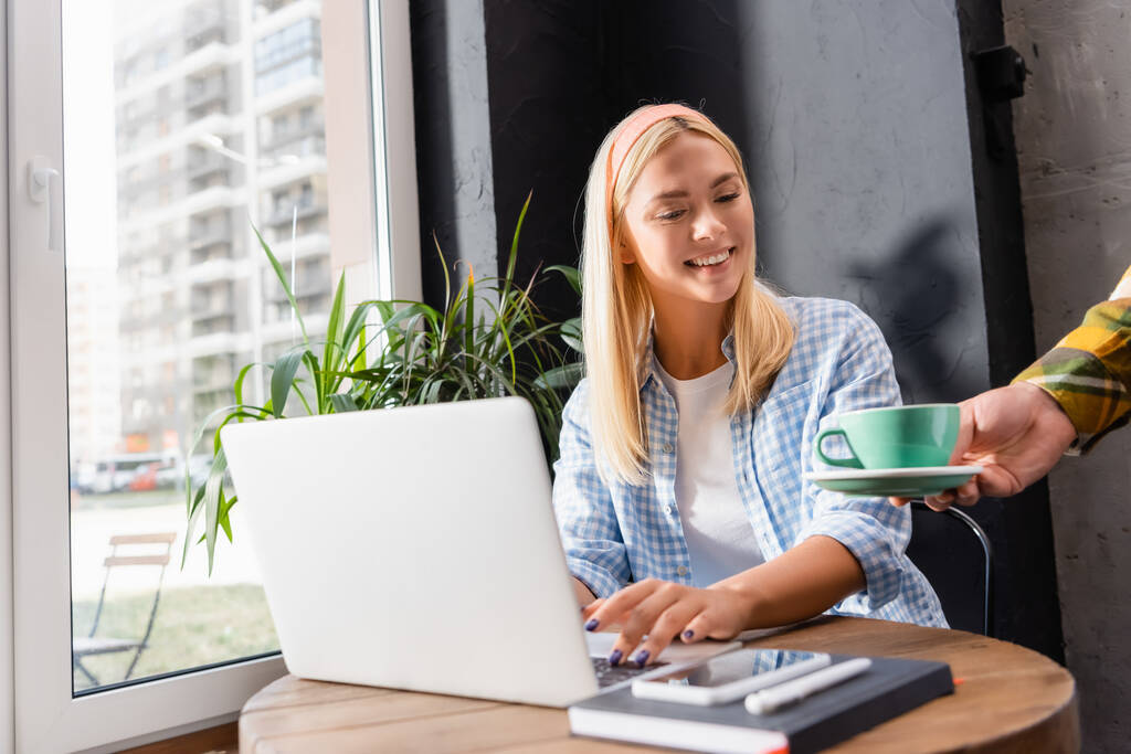 freelancer sorrindo digitando no laptop enquanto garçom servindo café, foreground borrado - Foto, Imagem