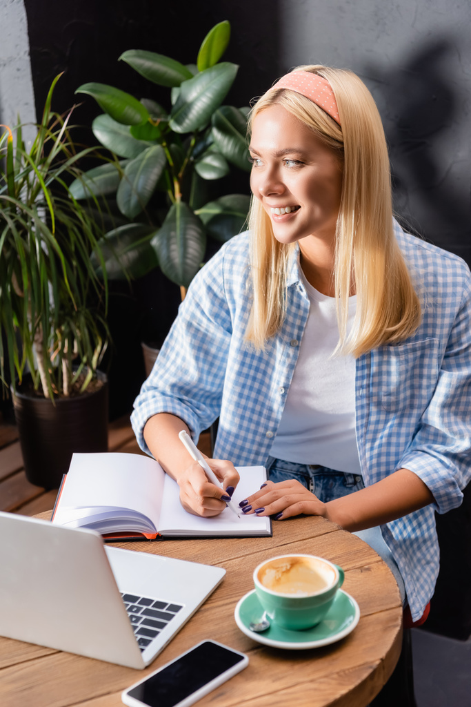 smiling blonde freelancer writing in notebook near laptop, cup of coffee and smartphone with blank screen in cafe - Photo, Image