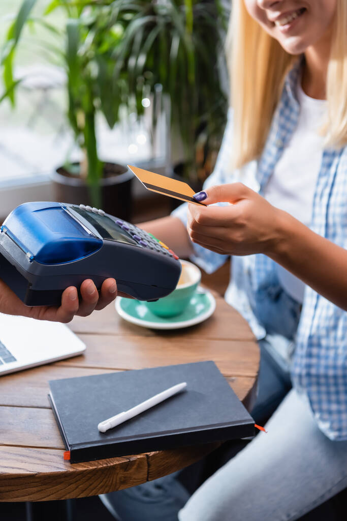 częściowy widok kelnera trzymającego terminal płatniczy w pobliżu uśmiechniętego freelancera z kartą kredytową na rozmytym tle - Zdjęcie, obraz
