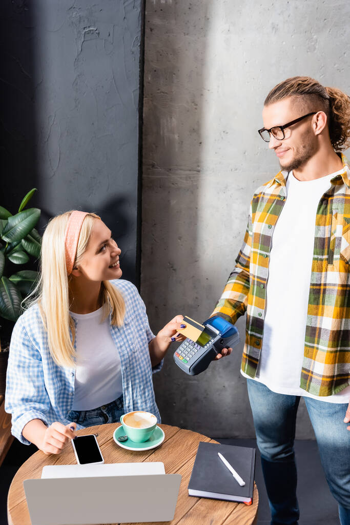 garçom sorrindo segurando terminal de pagamento perto de freelancer loira sentado no café perto de gadgets - Foto, Imagem