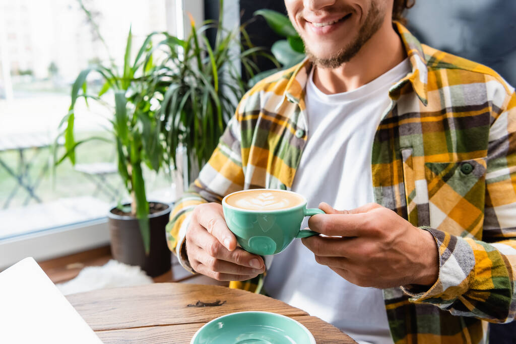 częściowy widok uśmiechniętego mężczyzny siedzącego w kawiarni i trzymającego filiżankę kawy z latte art - Zdjęcie, obraz