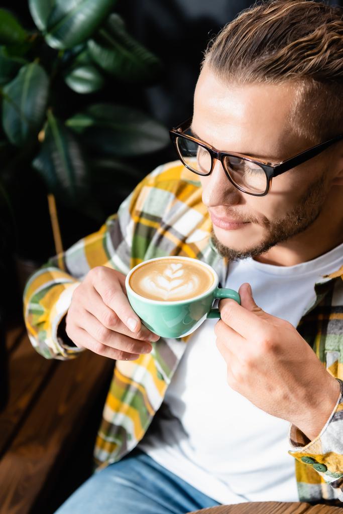 Blick aus der Vogelperspektive auf einen jungen Mann in Brille, der eine Tasse Kaffee mit Latte Art hält - Foto, Bild