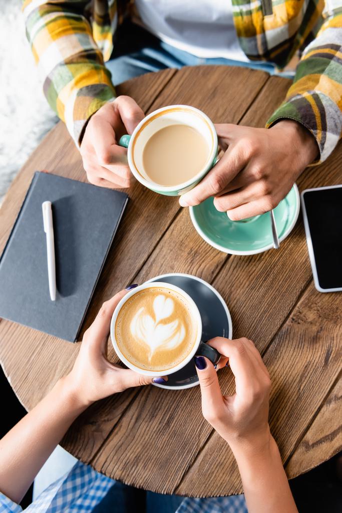 vista superior da mulher segurando xícara de café com arte latte perto de amigo no café - Foto, Imagem