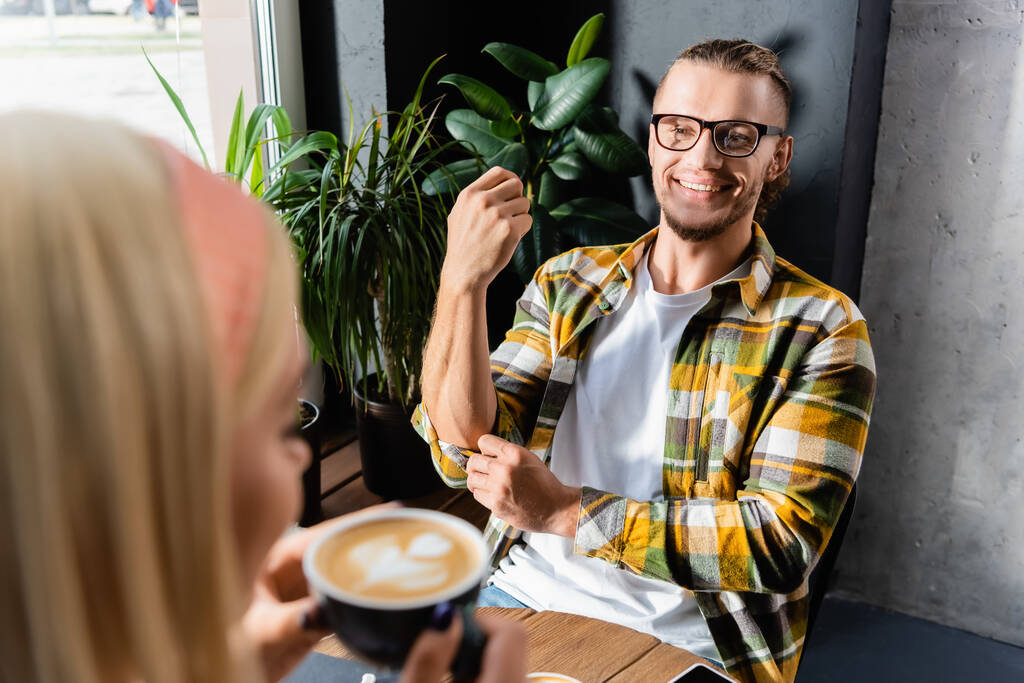 smiling young man in eyeglasses looking at woman holding cup of coffee on blurred foreground - Photo, Image