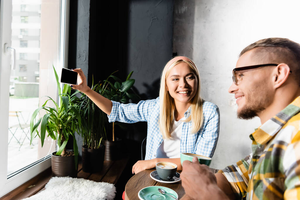 onnellinen blondi nainen ottaa selfie älypuhelimeen hymyilevä mies tilalla kahvikuppi hämärtynyt etualalla - Valokuva, kuva