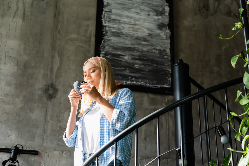 junge blonde Frau im karierten Hemd genießt den Geschmack von Kaffee, während sie eine Tasse auf der Treppe im Café hält - Foto, Bild