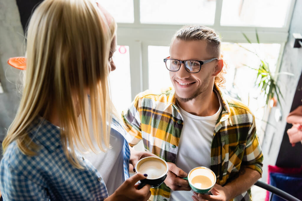 smiling young man in eyeglasses and woman on blurred foreground holding cups of coffee in cafe - Photo, Image