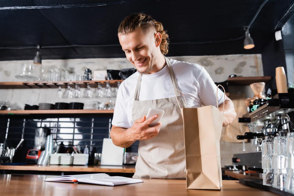 smiling barista in apron looking in notebook while holding smartphone near paper bag on bar counter - Photo, Image