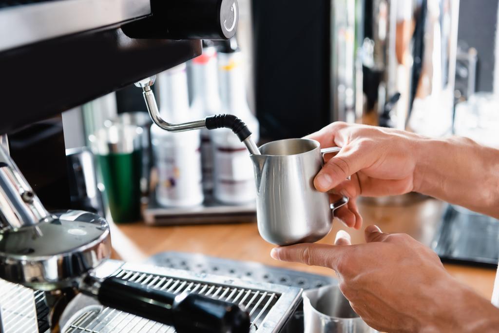 Kahve makinesinin yanında elinde metalik süt bardağı tutan baristanın kırpılmış görüntüsü. - Fotoğraf, Görsel