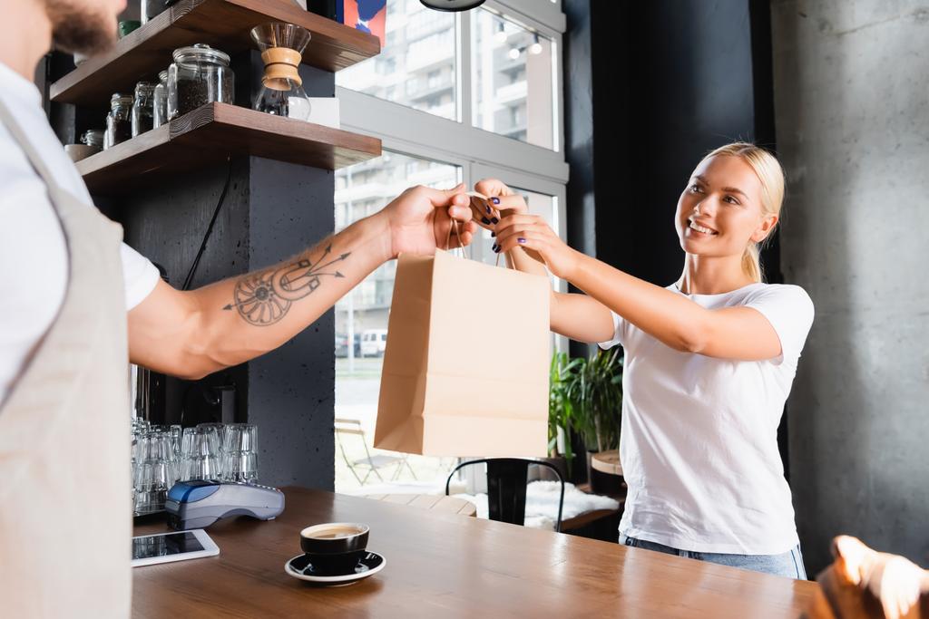 smiling blonde woman taking paper bag from tattooed barista on blurred foreground - Photo, Image