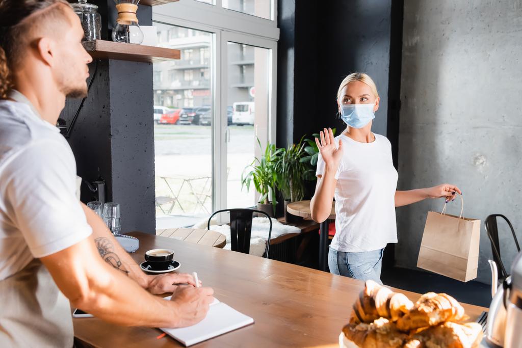 mladá žena v lékařské masce drží papírový sáček a mává rukou barista na rozmazané popředí - Fotografie, Obrázek
