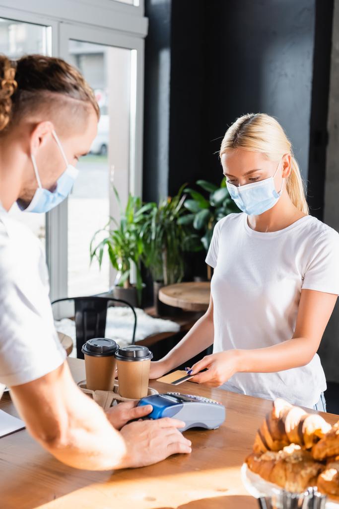 woman in medical mask paying for coffee to go through payment terminal near barista on blurred foreground - Photo, Image