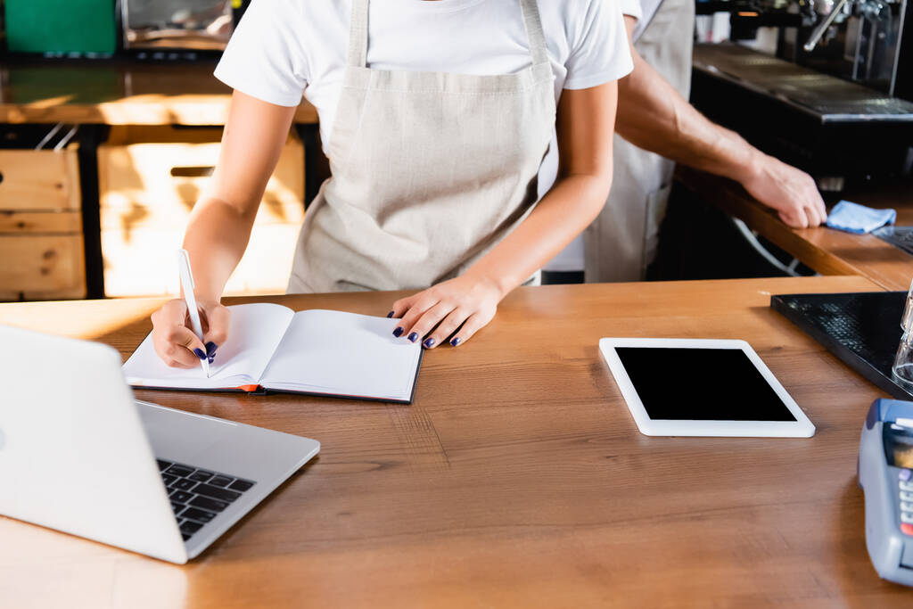 vista cortada da escrita barista em notebook perto de laptop, tablet digital com tela em branco e colega no fundo - Foto, Imagem