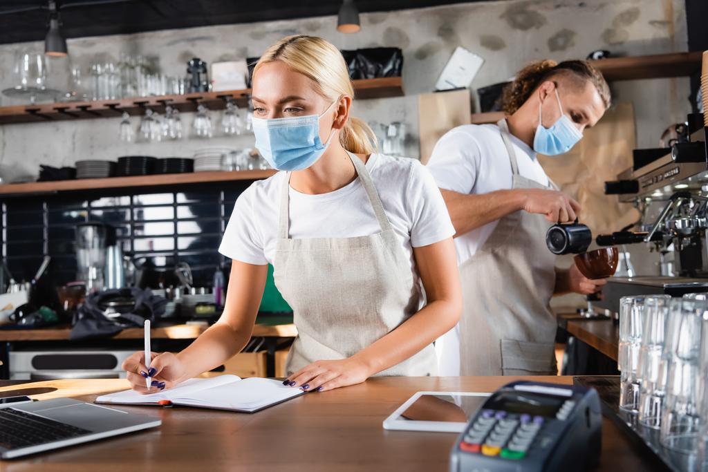 νεαρός σερβιτόρος στην ιατρική μάσκα γράφοντας στο σημειωματάριο κοντά στο laptop, και barista εργάζονται στο παρασκήνιο - Φωτογραφία, εικόνα