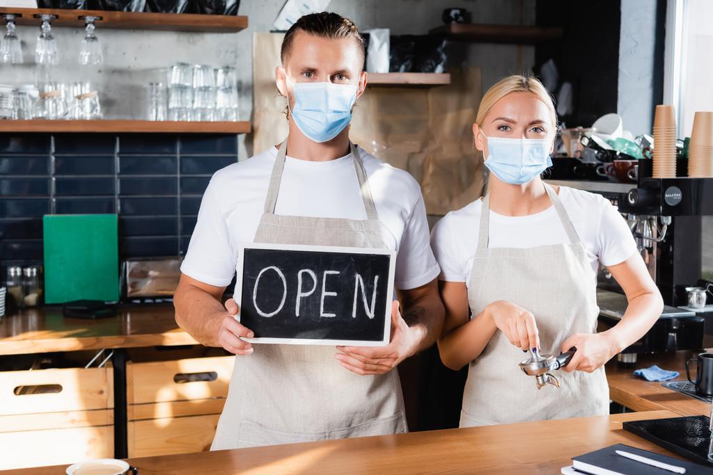 jong barista in medisch masker holding board met open letters in de buurt van collega met portafilter - Foto, afbeelding