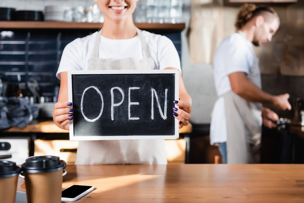 vista cortada de garçom sorridente segurando placa com letras abertas perto barista trabalhando em fundo borrado - Foto, Imagem