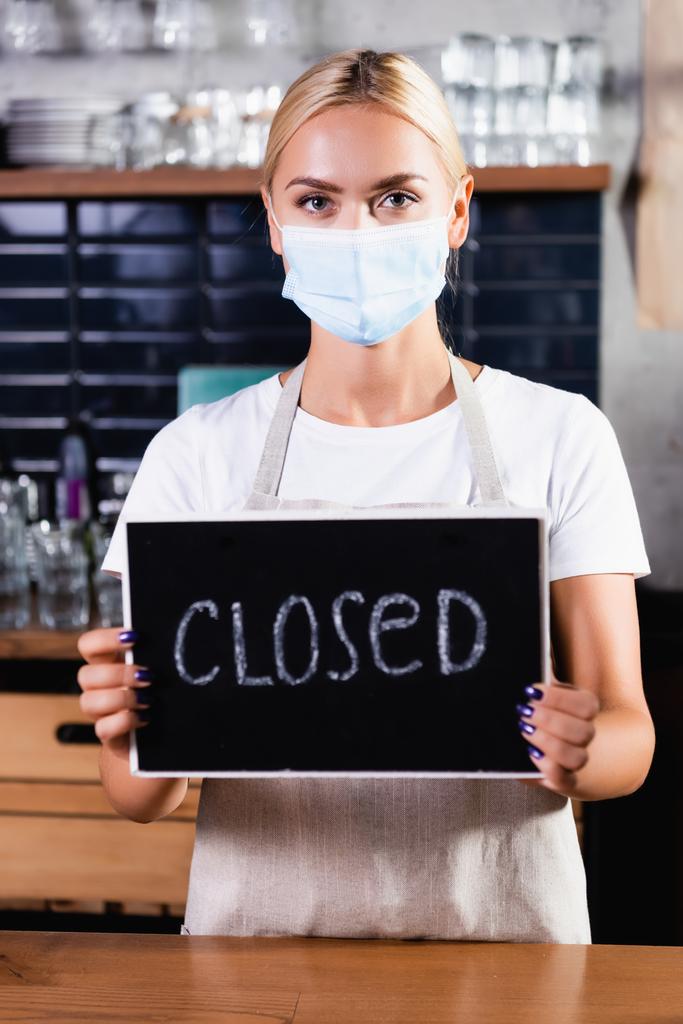 blond barista in medisch masker holding board met gesloten belettering in cafe - Foto, afbeelding