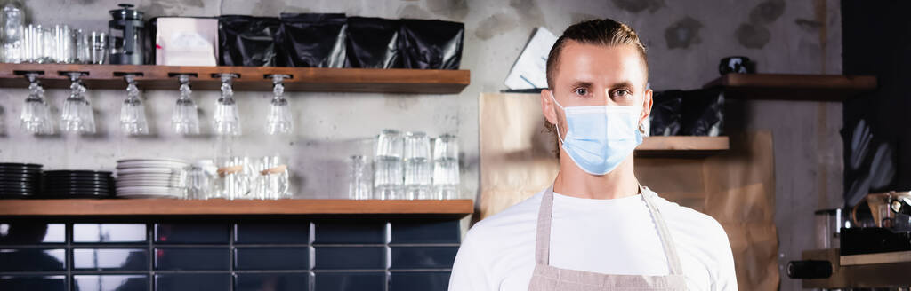 бариста в медицинской маске смотрит в камеру, стоя на рабочем месте в кафе, баннер - Фото, изображение