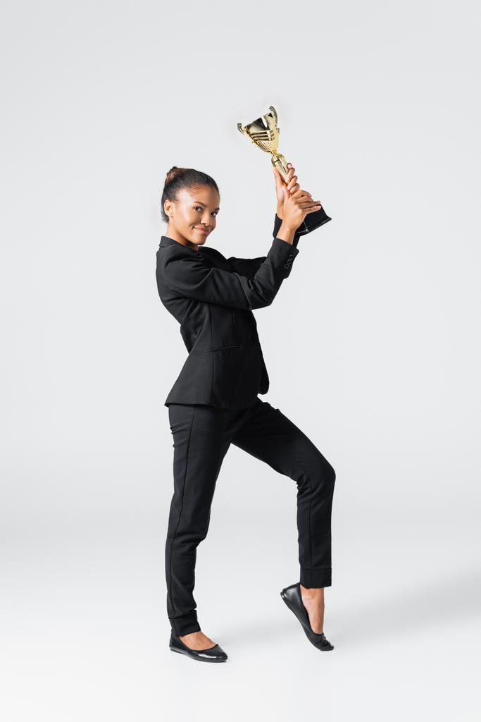 sonriente mujer de negocios afroamericana con copa de oro aislada en gris - Foto, Imagen