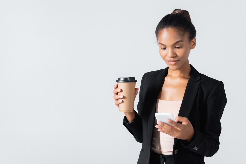 mulher de negócios afro-americana com copo de papel usando smartphone isolado em cinza - Foto, Imagem