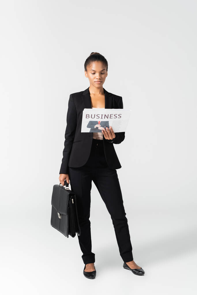 Αφροαμερικανή επιχειρηματίας με βαλίτσα ανάγνωση επιχειρηματική εφημερίδα απομονωμένη σε γκρι - Φωτογραφία, εικόνα