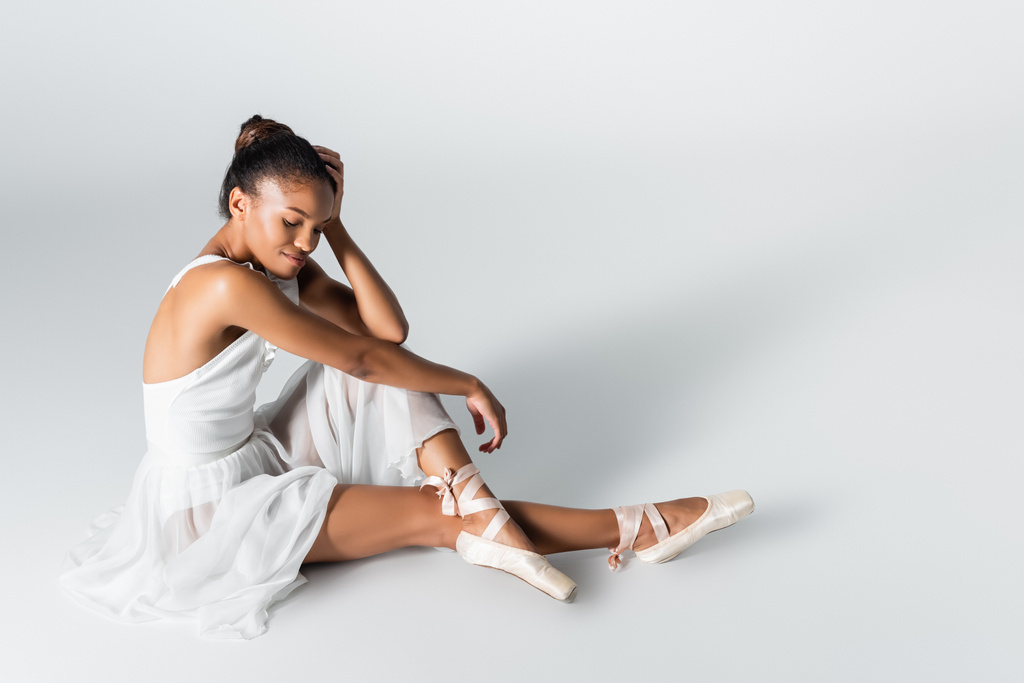 anmutige afrikanisch-amerikanische Ballerina im Kleid, sitzend auf dem Boden auf weißem Hintergrund - Foto, Bild