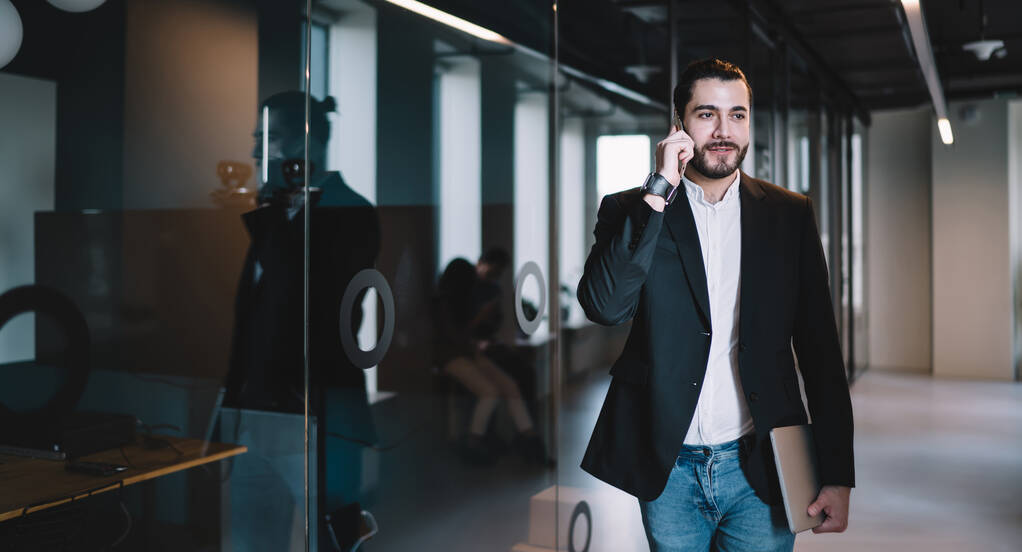 Moderno uomo d'affari con blazer nero e jeans che camminano nel corridoio dell'ufficio con il computer portatile in mano e discutono di affari per telefono - Foto, immagini