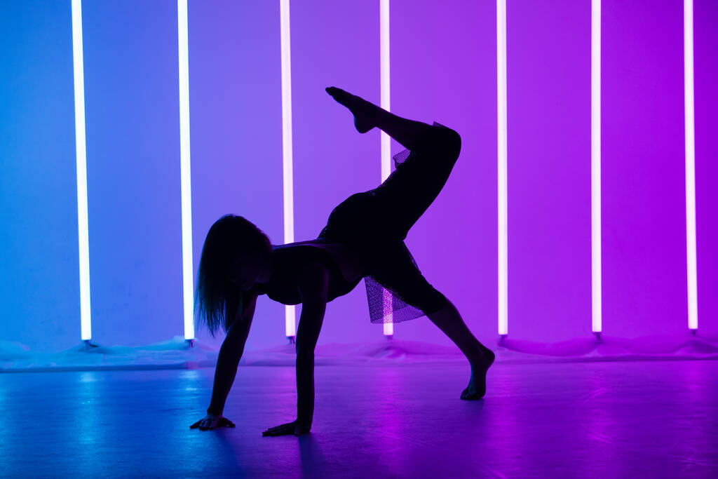 Silueta mladé tanečnice tančící a pózující v místnosti s neonovými lampami. Štíhlý moderní model v krásné póze na fialovém modrém pozadí. - Fotografie, Obrázek