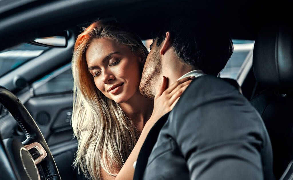 情熱的なカップル車の中で愛を作る。魅力的なブロンドでエレガントな黒ドレスとハンサムなビジネスマンでスーツあります接吻で車. - 写真・画像