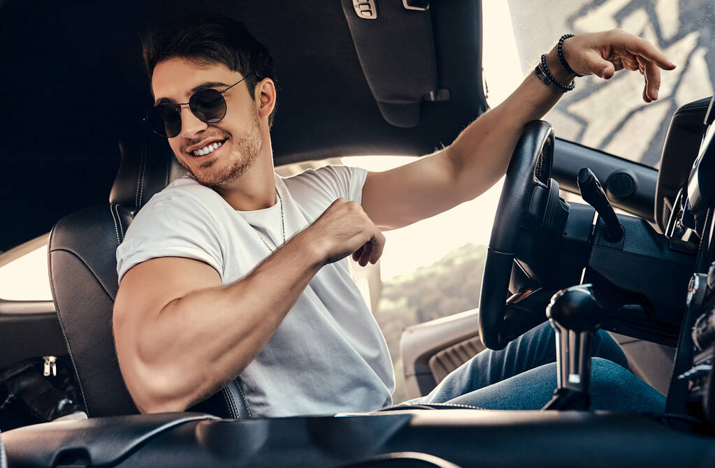 Jóképű fiatalember napszemüvegben és alkalmi ruhában ül a modern sportkocsijában. - Fotó, kép