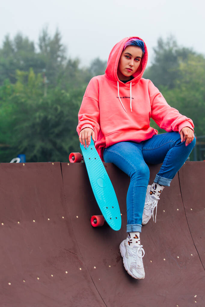 Summer lifestyle image of trendy pretty young girl dressed in pink hoodie and jeans sitting next to the skateboard court with her blue plastic skateboard. - Photo, Image