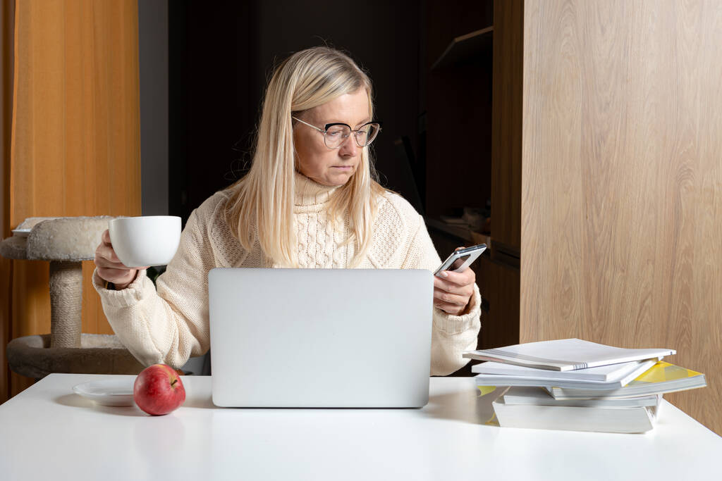 blondi keski-ikäinen liikenainen istuu pöydän ääressä juomassa teetä ja työskentelemässä kannettavan tietokoneen kanssa kotitoimistosta - Valokuva, kuva