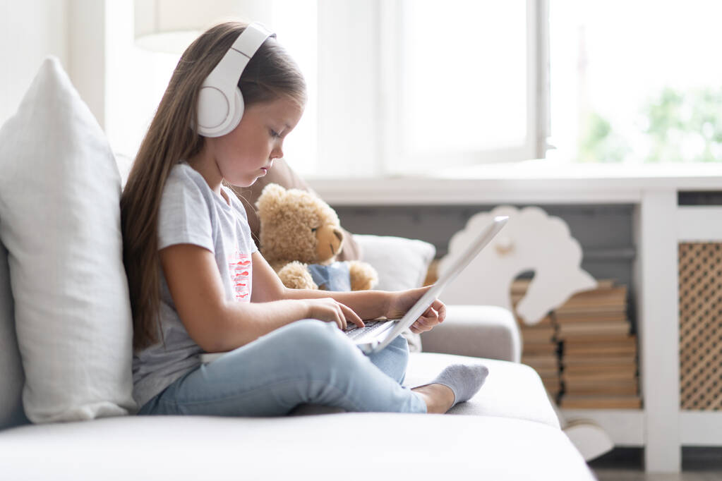 Online-Bildung. Süßes Mädchen mit Kopfhörern auf der Suche nach Videolehrerkonferenz Laptop sitzt auf der Couch zu Hause - Foto, Bild