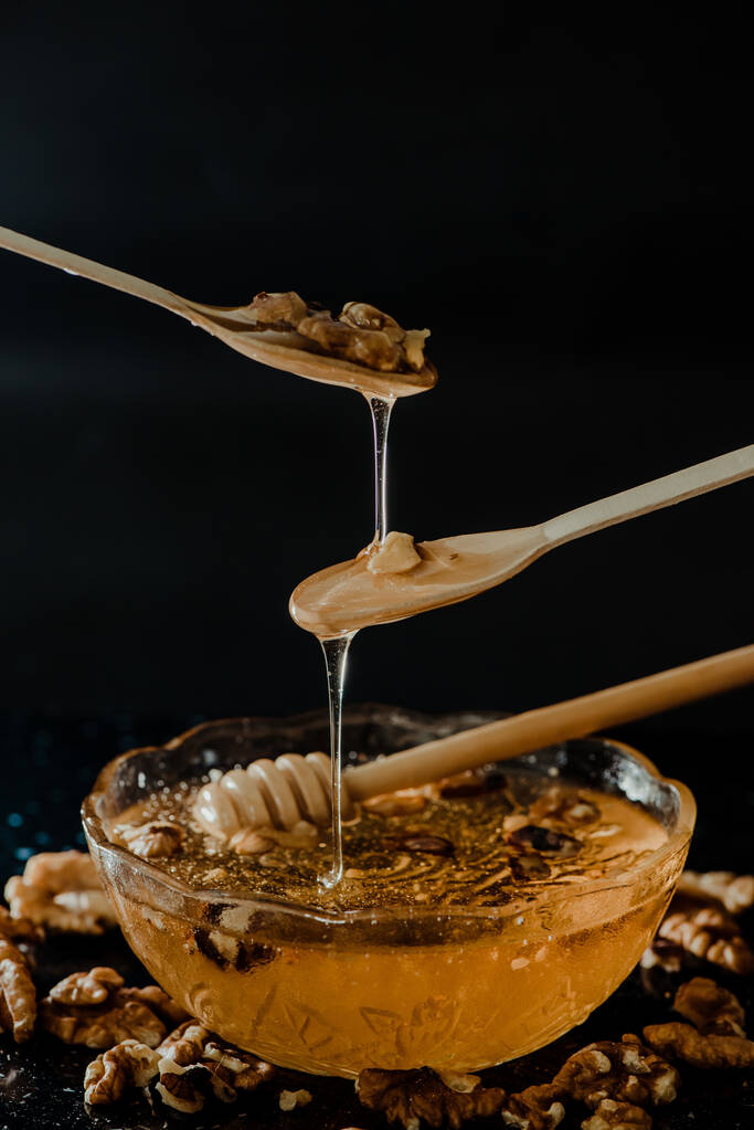 Honiglöffel aus Holz, eingetaucht in ein Glas voller Honig und Nüsse auf schwarzem Hintergrund. Honig tropft herum, ansprechende und einladende Fotos. - Foto, Bild