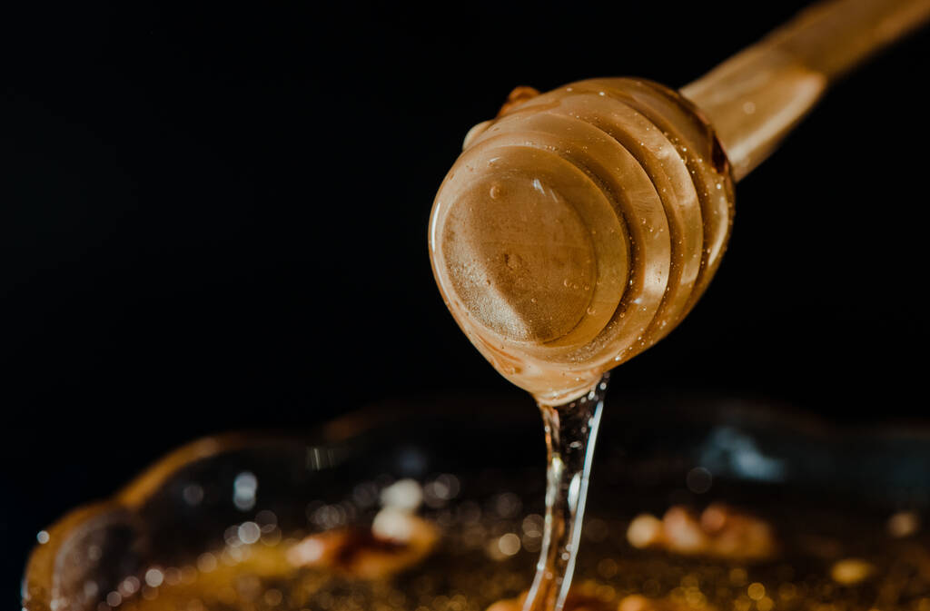 Honiglöffel aus Holz, eingetaucht in ein Glas voller Honig und Nüsse auf schwarzem Hintergrund. Honig tropft herum, ansprechende und einladende Fotos. - Foto, Bild