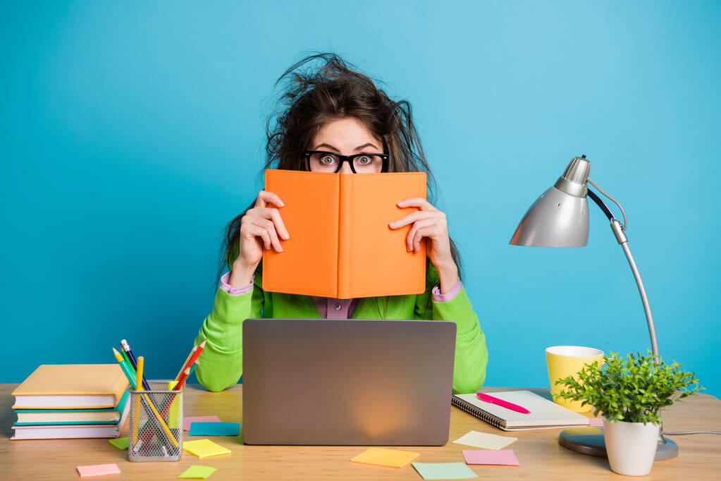Retrato de asombrado marrón desordenado cabello chica sentarse escritorio cerrar cubierta libro cara aislado color azul fondo - Foto, Imagen