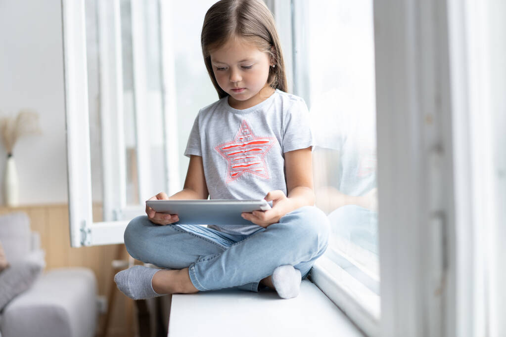 Cute małe dziecko za pomocą tabletu cyfrowego w domu wnętrza z dużym oknem. - Zdjęcie, obraz