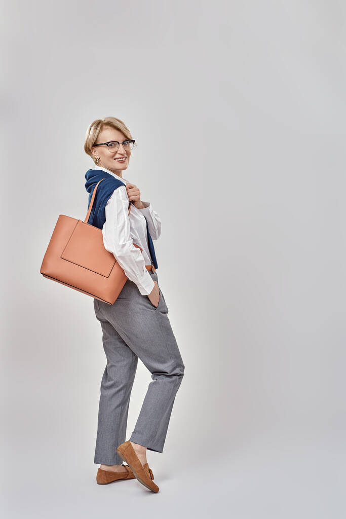 エレガントな中年の白人女性がビジネス服を着てカメラで笑顔しながらハンドバッグを振り、灰色の背景の上に隔離された立って - 写真・画像