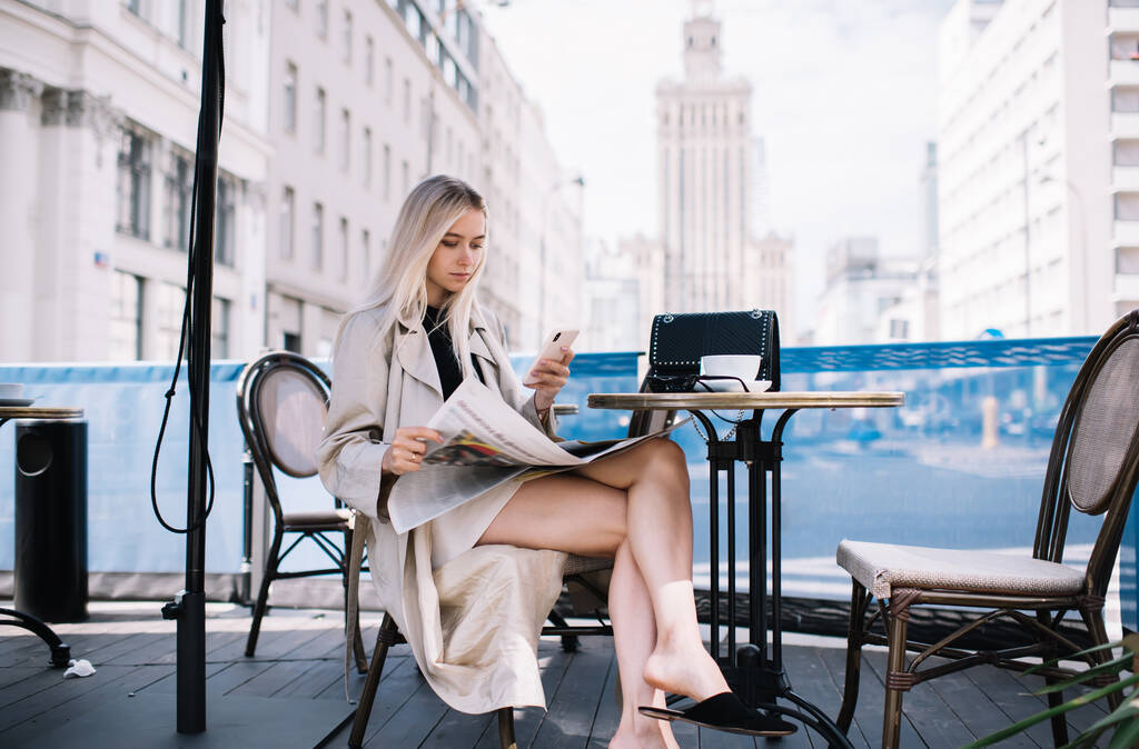 Модна жінка використовує смартфон у впевненій позі, проводячи час з газетою і чашкою кави в тротуарному кафе вдень
 - Фото, зображення
