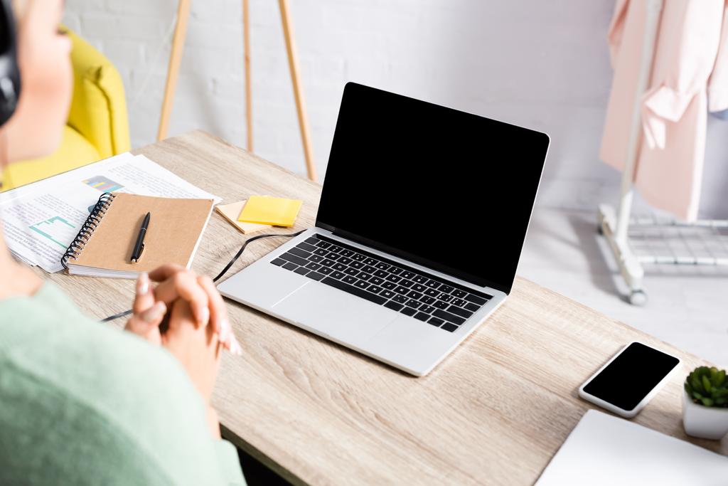 Ноутбук, смартфон и бумаги на столе рядом с телеработником в наушниках на размытом переднем плане  - Фото, изображение