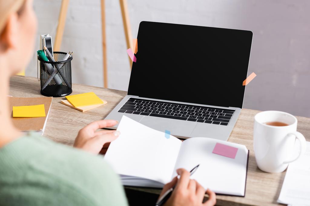 Laptop com tela em branco perto de notas pegajosas, xícara de chá e freelancer segurando caneta e notebook em primeiro plano desfocado  - Foto, Imagem