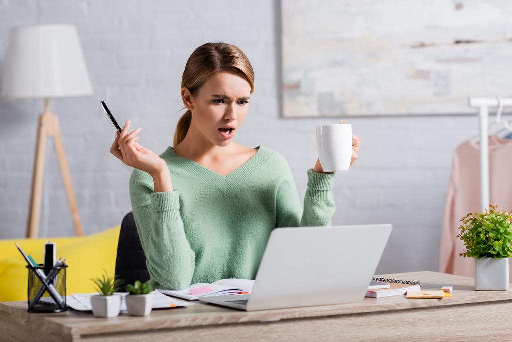 Freelancer confuso sosteniendo la pluma y la taza mientras mira el portátil cerca de los cuadernos en primer plano borroso  - Foto, imagen