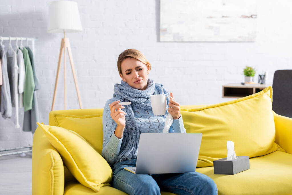 Chora kobieta trzyma termometr i kubek w pobliżu serwetek i laptopa na kanapie  - Zdjęcie, obraz